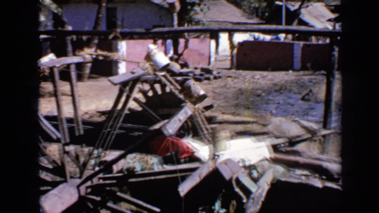Muddy flooded farm area with wooden debris, poles, and low houses in the background.