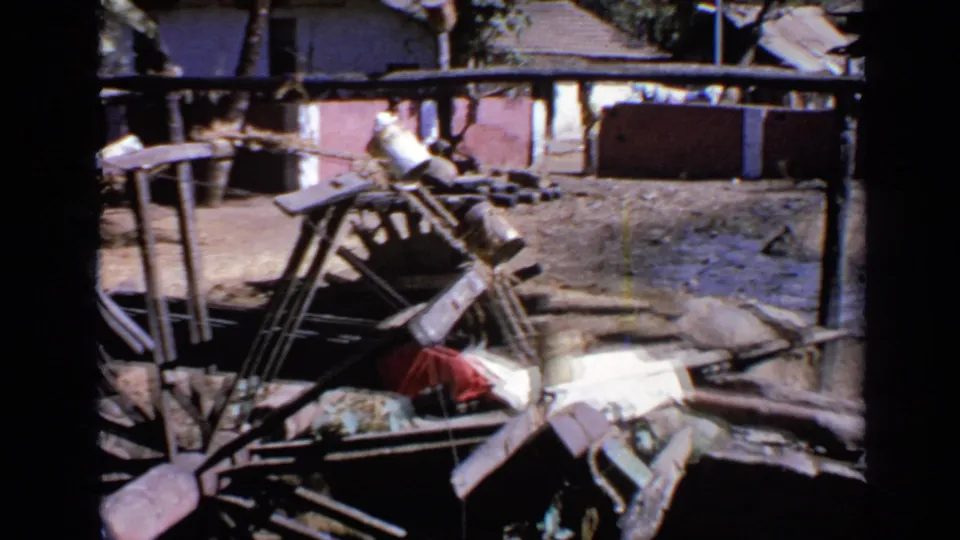 1970 India 8mm frame showing a flooded rural farm area with scattered wood, poles, bare earth, and low tiled-roof houses behind.