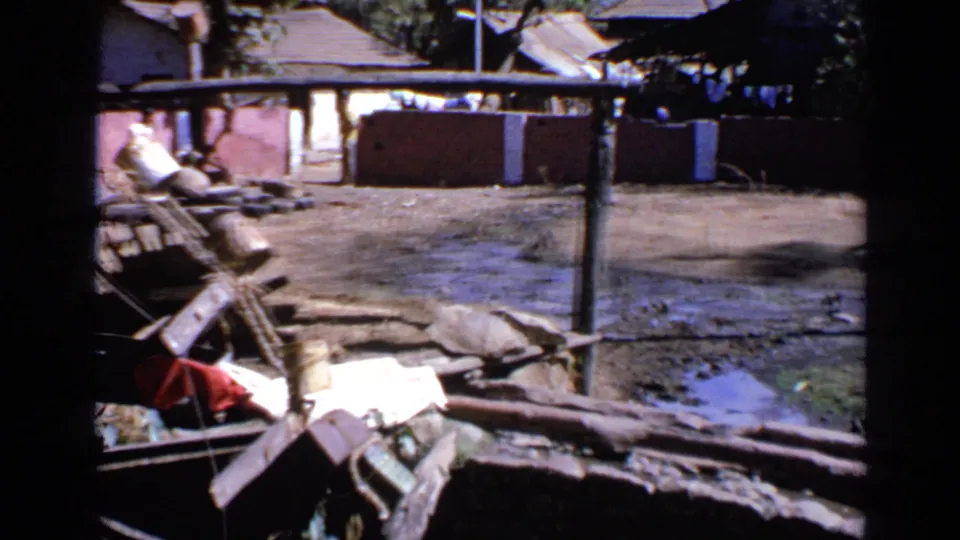 1970 India home-movie frame of a muddy flooded rural yard with timber debris in front of simple low buildings and trees.