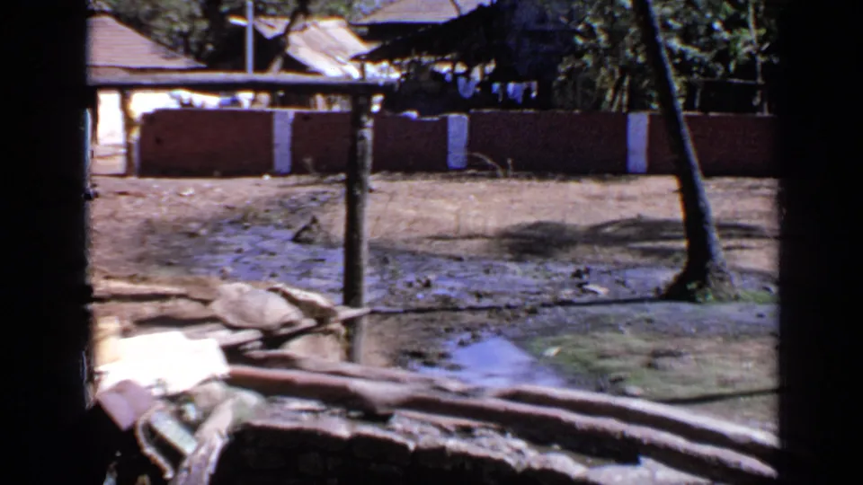 1970 India 8mm frame showing pooled floodwater, muddy ground, a low wall, roofed village houses, and a leaning palm tree.
