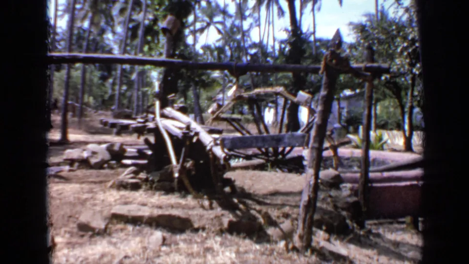 1970 India home-movie frame with rough wooden supports, cart-like parts, dry soil, palms, foliage, and a purple barrel-like container at the side.