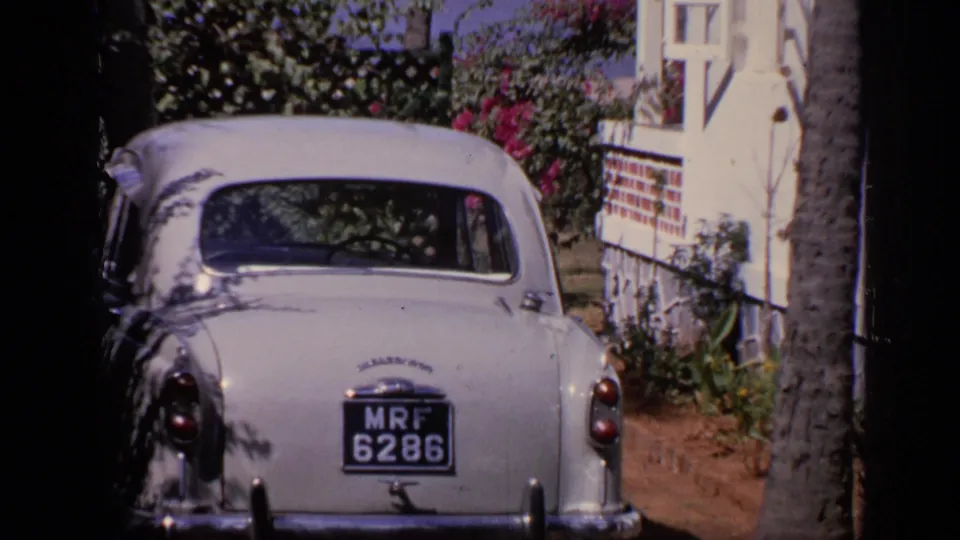 White Ambassador car parked near a colonial-style house in India, with greenery and flowers around the driveway.