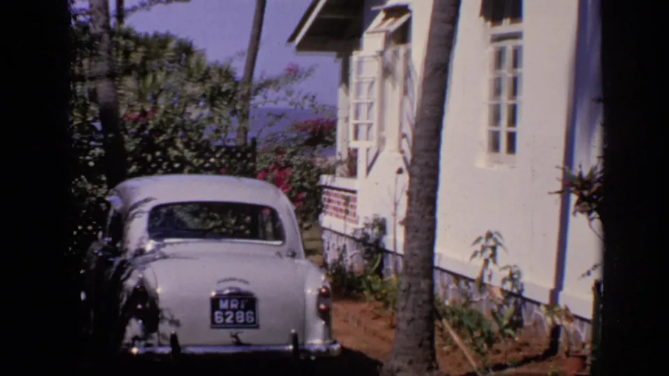 Wider 1970 India home-movie frame showing the white Ambassador by a house, with palm trunk, windows, and garden planting.