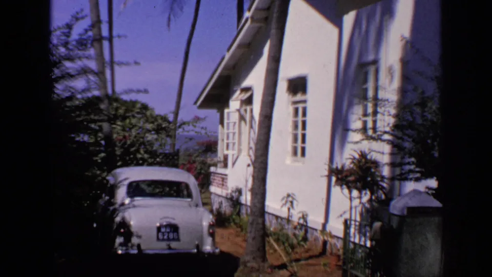 Wide 8mm frame from India showing the car at the end of a garden path beside a white house and tall palms.