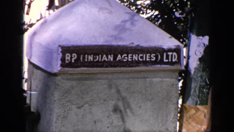 Close 8mm frame of a BP (Indian Agencies) Ltd sign on a pale concrete pillar, with trees and shadowed edges around the frame.