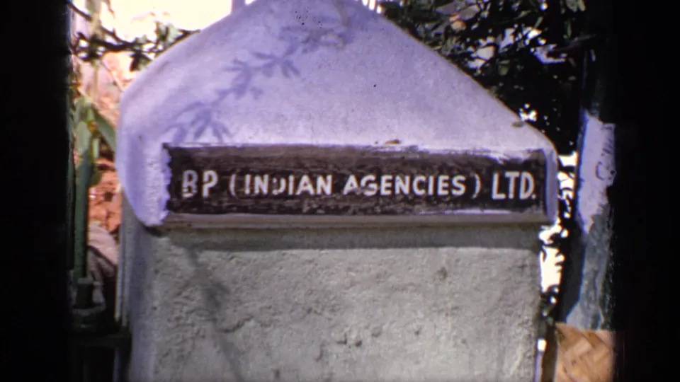 1970 India 8mm frame showing a concrete pillar with a BP Indian Agencies Ltd. sign, partly shaded by leaves.