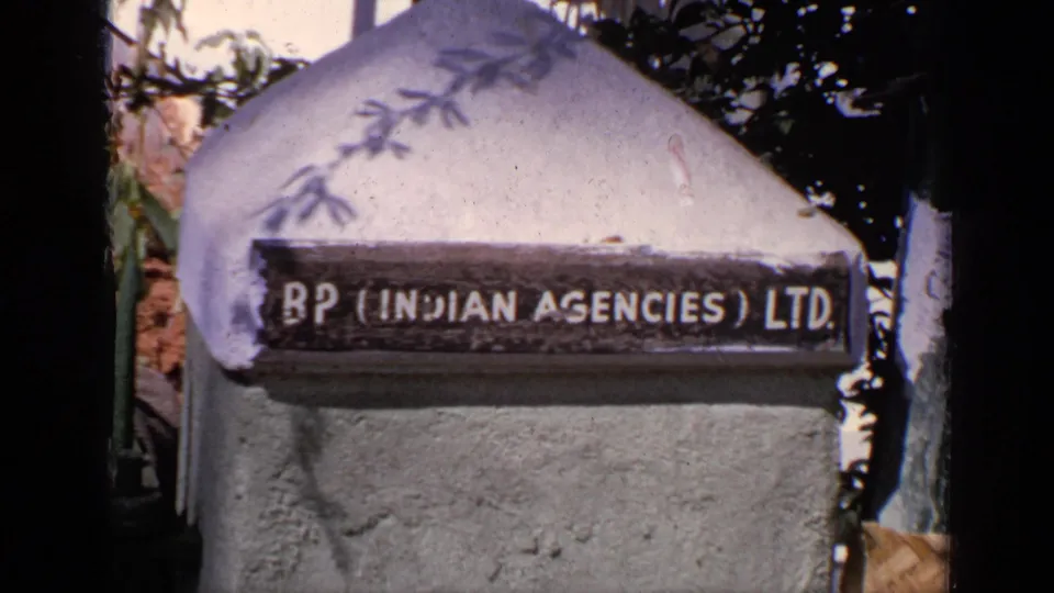 1970 India 8mm frame with a weathered BP Indian Agencies Ltd. sign mounted on a concrete pillar near trees.