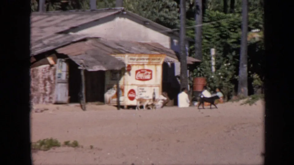 1970 India 8mm frame showing a rustic roadside building, Coca-Cola cart, seated people, and animals near palm trees.