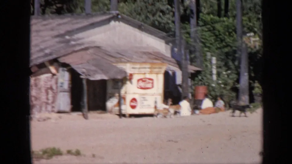 1970 India 8mm frame of a Coca-Cola cart at a dusty roadside stop with seated people, animals, palms, and greenery.