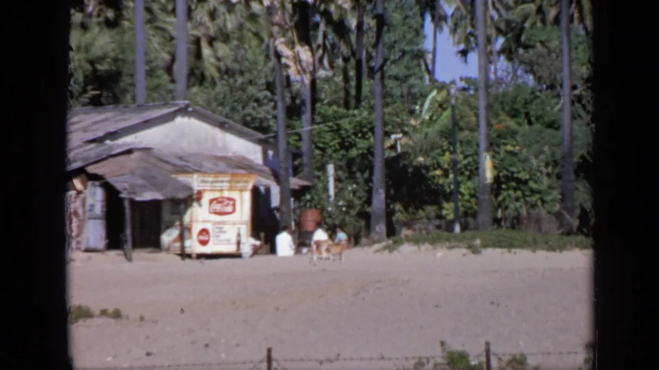 Close 1970 8mm view in India of a palm-backed beach shack with a Coca-Cola sign, people beside it, and a dog crossing the sand.