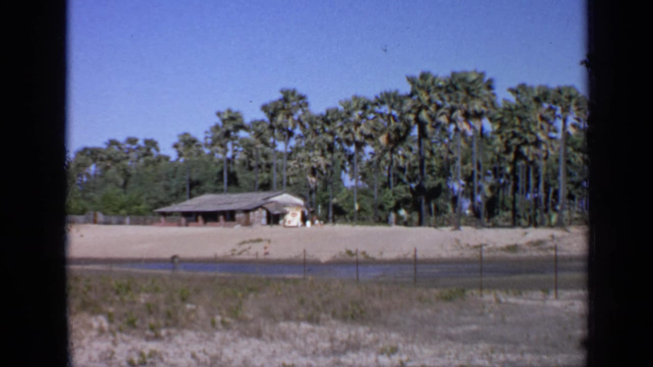 Low building beyond sandy ground, fence, waterway, and palm trees under blue sky.