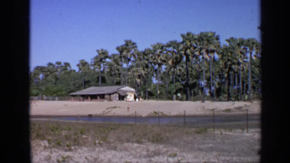 1970 India 8mm view of a weathered low building across a sandy open area, with a water channel, fence posts, and palms behind it.