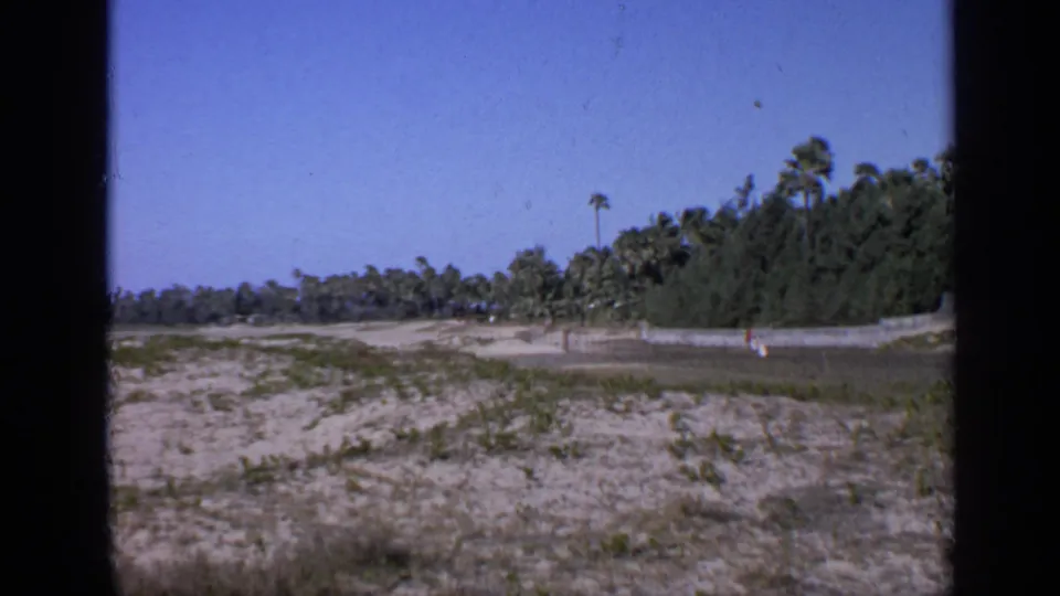 1970 India 8mm frame looking along sandy ground and a fence line toward palms, scrub vegetation, and distant low structures.