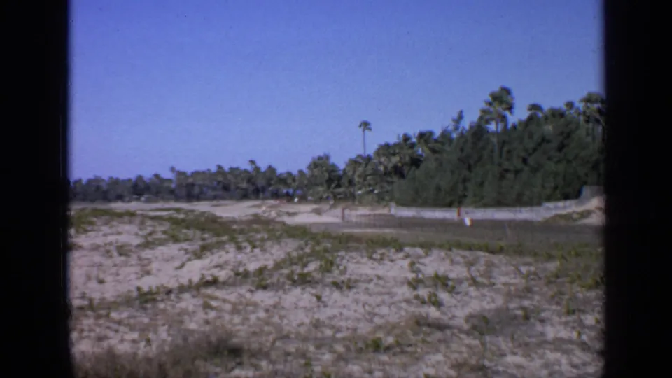 1970 India 8mm frame with a wide sandy lot, scattered plants, palm trees, and a low wall in the distance.