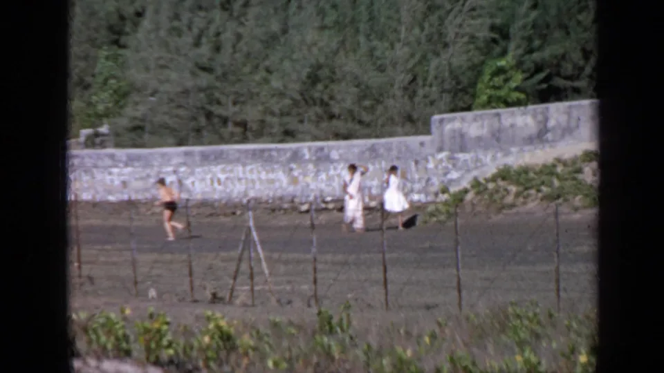 1970 India 8mm frame showing a runner crossing a dirt track as two women in white walk beside a stone wall.