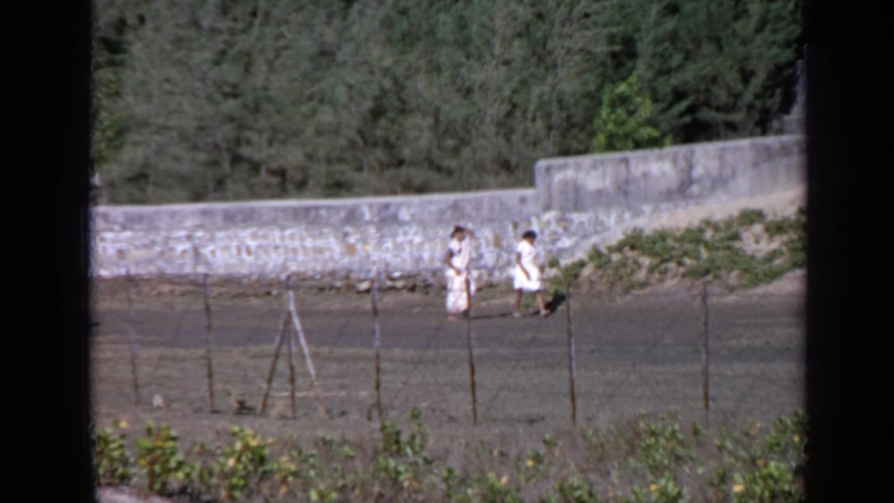 Two people in light clothing walk beside a stone wall on a dirt path behind a wire fence.