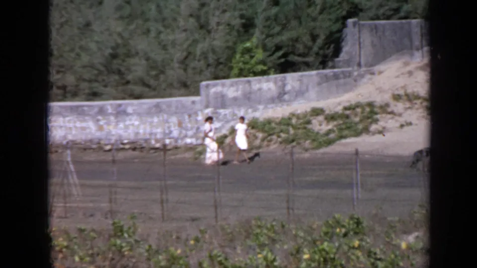 Two figures walk beside a curved stone wall in India, filmed from a distance on 8mm with a wire fence in the foreground.