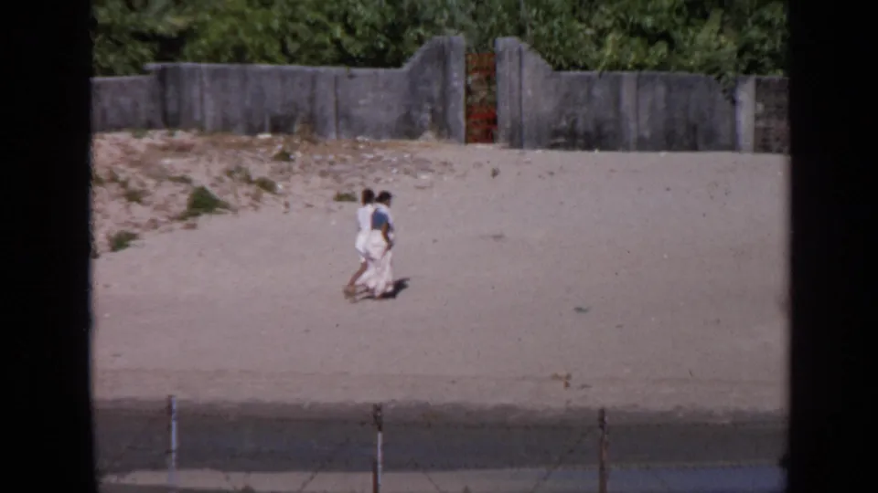 Two adults walk side by side across a sunlit sandy embankment in India, with a concrete wall and foliage behind them.