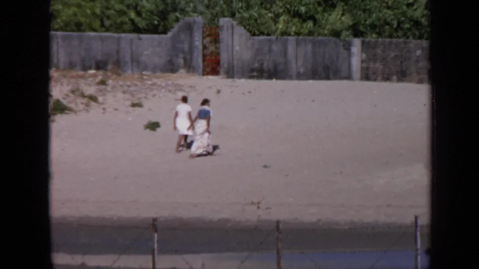The couple continues across the sandy ground, seen from a distance against a concrete wall and dense green foliage in 1970 India footage.