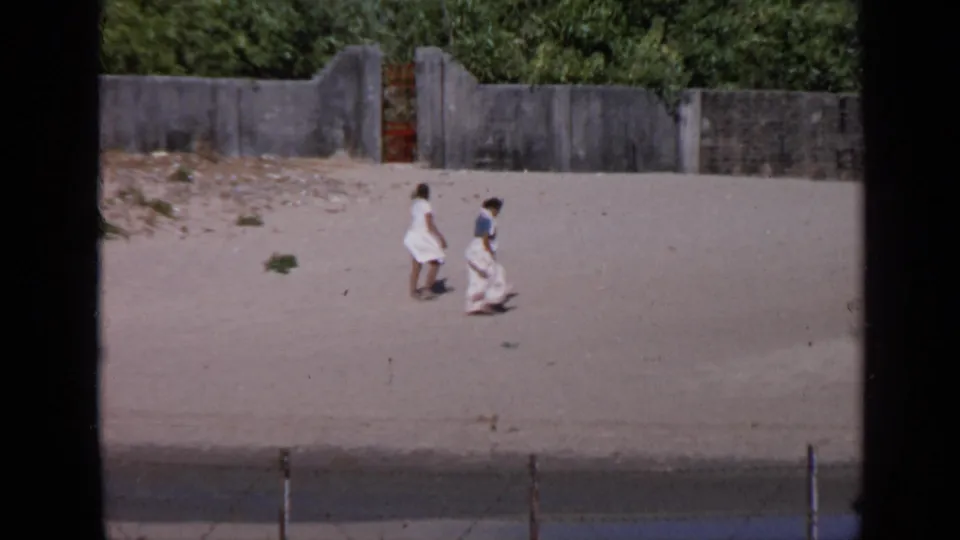 The couple walks farther across the sandy embankment beneath a weathered concrete wall, filmed from a distant home-movie viewpoint in India.