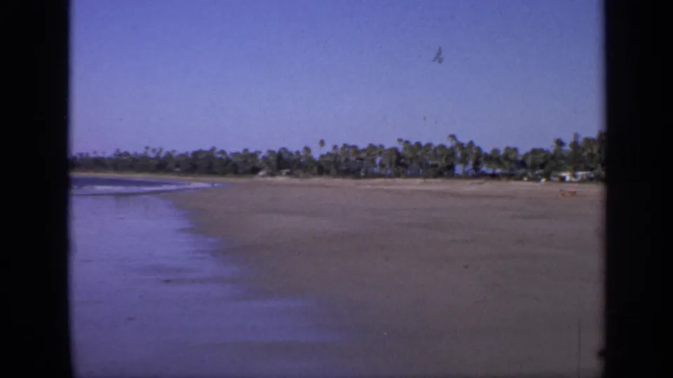 1970 India 8mm frame showing wet reflective sand, a narrow line of surf, distant palms, and a small figure on the beach.