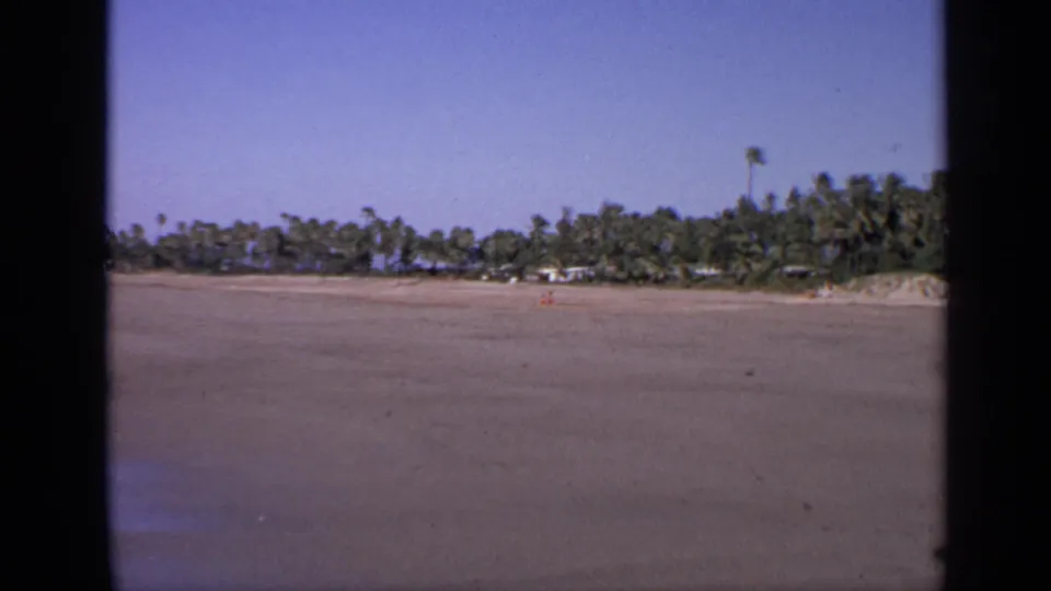 1970 India coastal home-movie frame showing a mostly empty sandy beach backed by palms and low greenery.