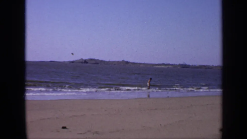 1970 India beach frame with one distant person in the surf, small waves, sand, blue sky, and low land on the horizon.