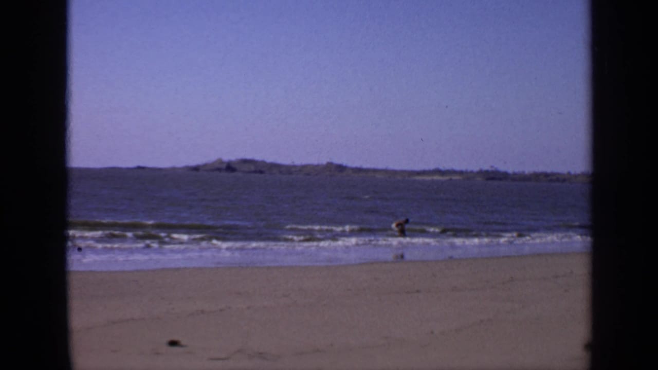 Man standing in shallow surf on a sandy beach with distant land across the water.