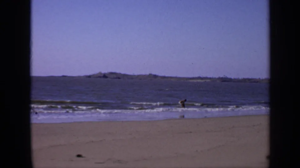 1970s India home-movie frame showing a young man near small waves on a sandy beach, with low distant land across the sea.