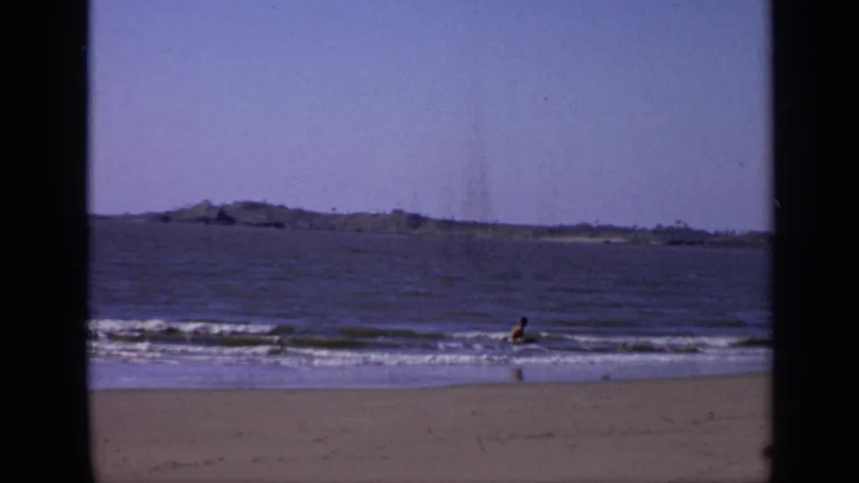 1970s India beach footage frame with a young man wading in gentle surf beneath a clear sky and distant shoreline.