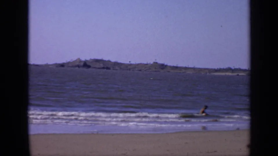 1970s India home-movie frame showing the man low in the shallow water, with beach sand in front and distant land across the sea.