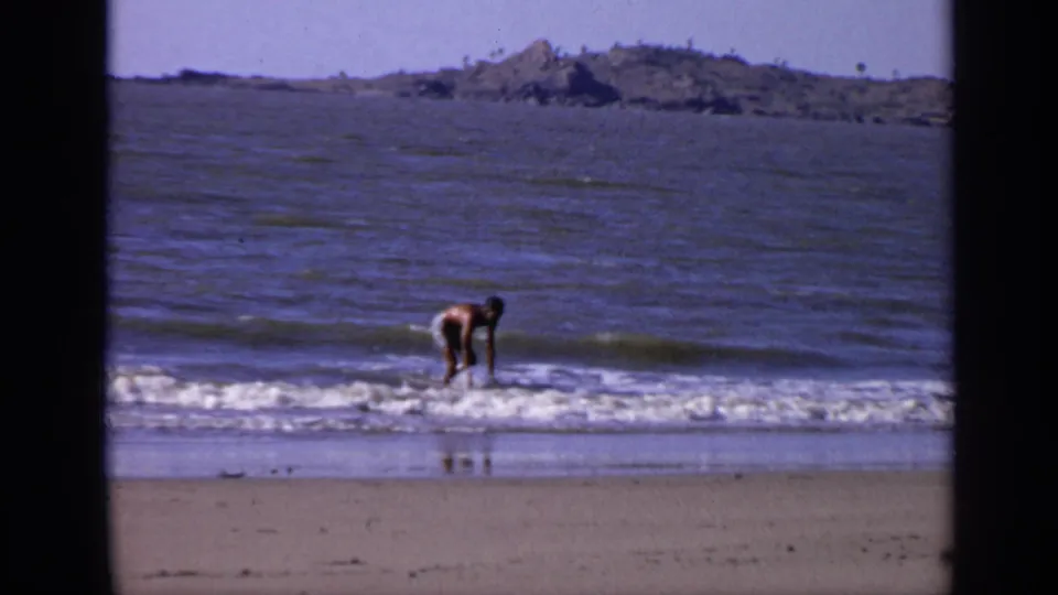 1970s India beach frame showing a young man bending in shallow waves, with calm sea and distant land in the background.
