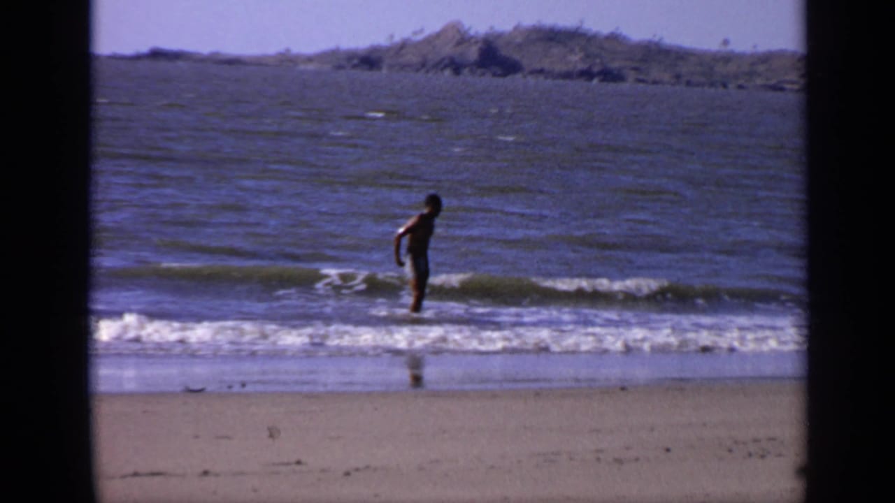 Man in swim trunks standing in shallow ocean waves beside a sandy beach.