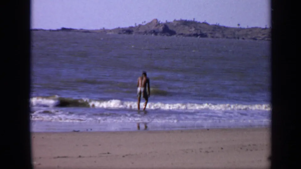 1970 India coastal frame with a man in swim trunks facing the camera from shallow waves, with sand and distant rocks visible.