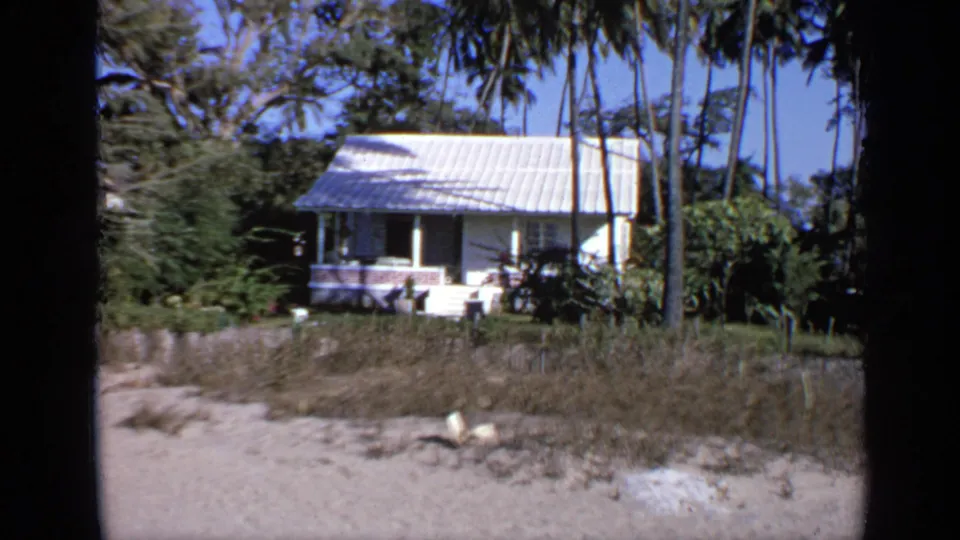 The 8mm frame shows the white rural house again, framed by palm trees, scrubby foreground grass, and bright blue sky.