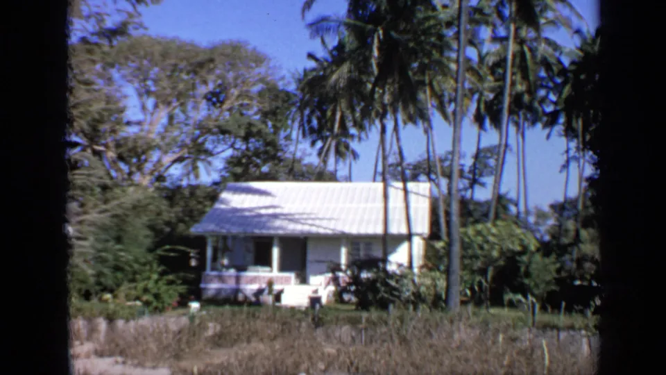 1970 India home-movie view of a small white house, palms, and garden foliage framed by dark vertical edges.
