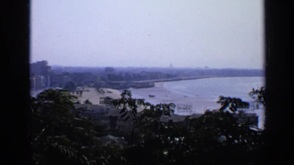 1970 India 8mm frame showing a hazy coastal city overlook with shoreline buildings, calm water, and dark foreground foliage.
