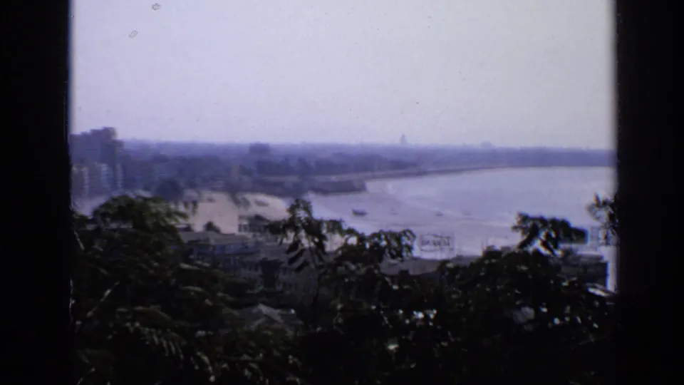 1970 India home-movie frame of a hazy waterfront cityscape, with beach, buildings, calm water, and silhouetted trees in front.