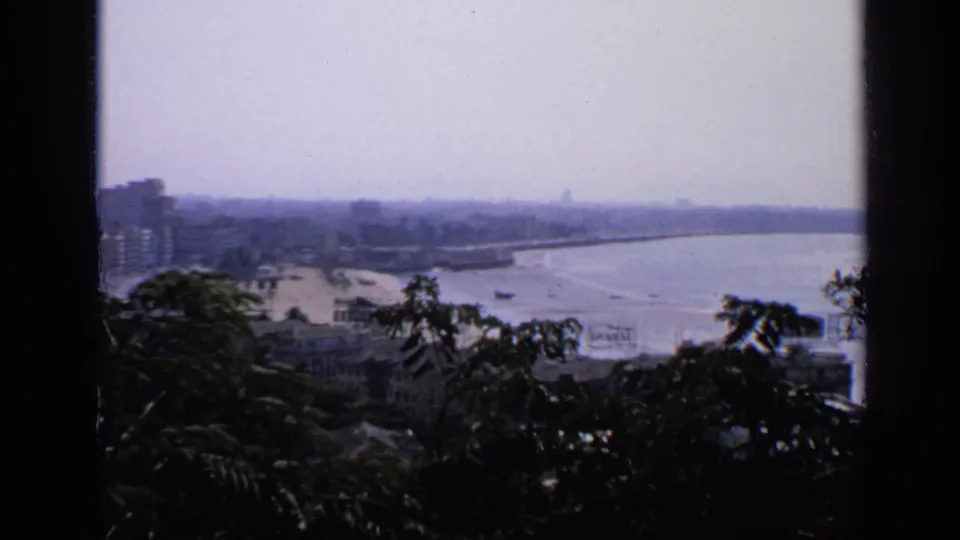 1970 8mm home-movie view in India showing a hazy coastline, sea, small boats, shoreline buildings, and silhouetted foliage.