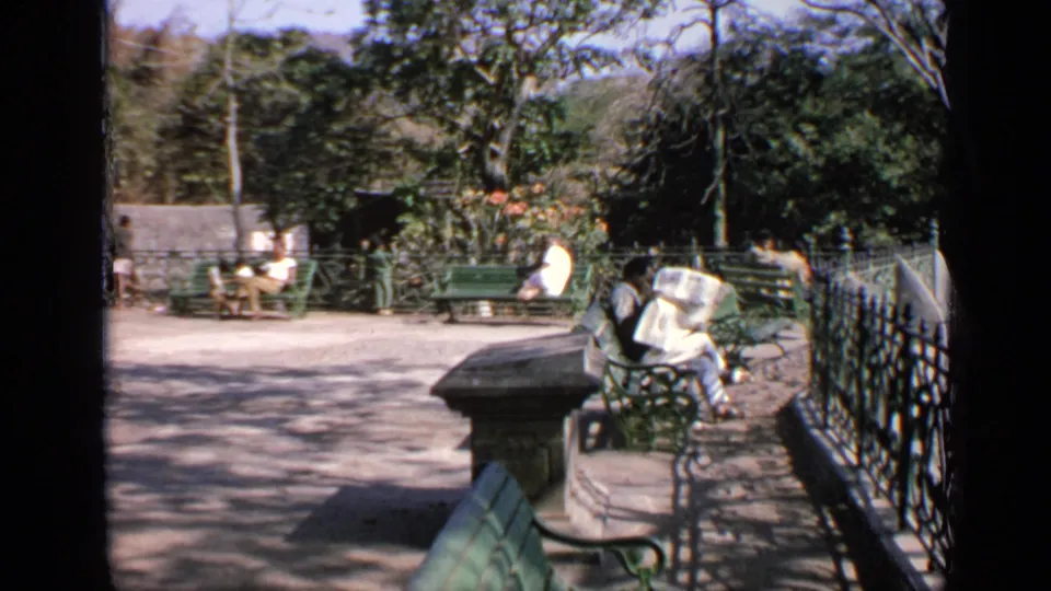 A sunny 1970 India park scene shows several people seated on green benches beside trees, flowers, a railing, and paved paths.