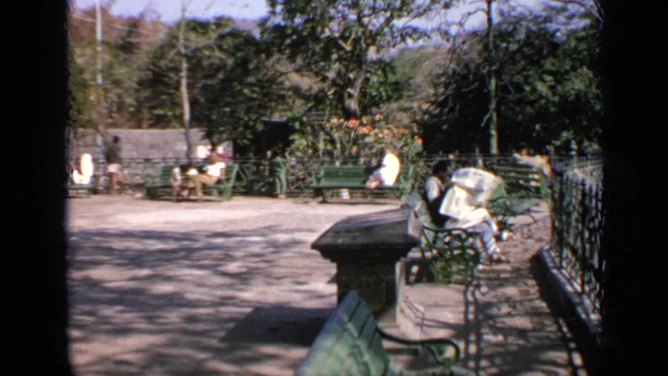 1970 India home-movie frame showing green benches, seated people, trees, and a paved public area.