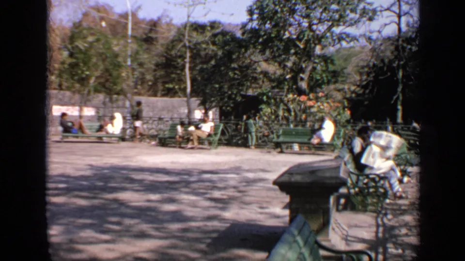 1970 India home-movie view of a public seating area with benches, trees, and distant pedestrians.
