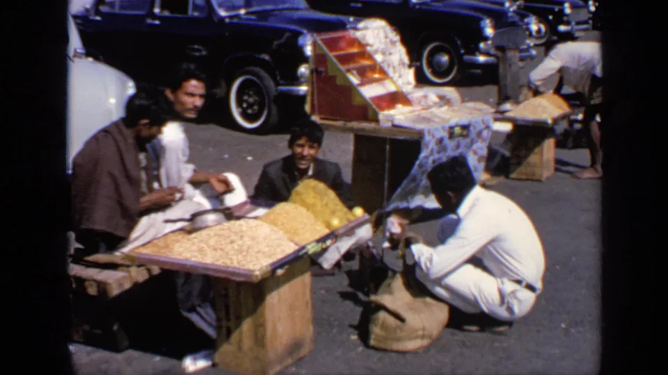 1970 India street-market frame showing vendors around wooden trays of goods with older cars in the background.