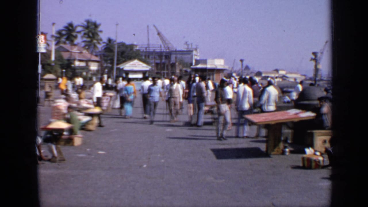 1970 India street scene with pedestrians walking past small roadside stalls, palm trees, older buildings, and construction cranes in the distance.