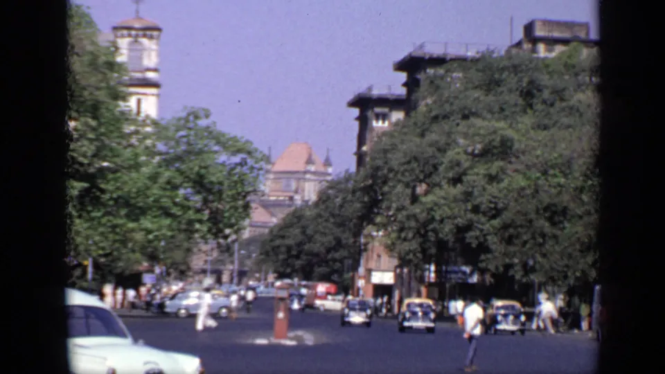 1970 India urban street view with older cars, pedestrians crossing, large trees, a tower, and multi-story buildings along the road.