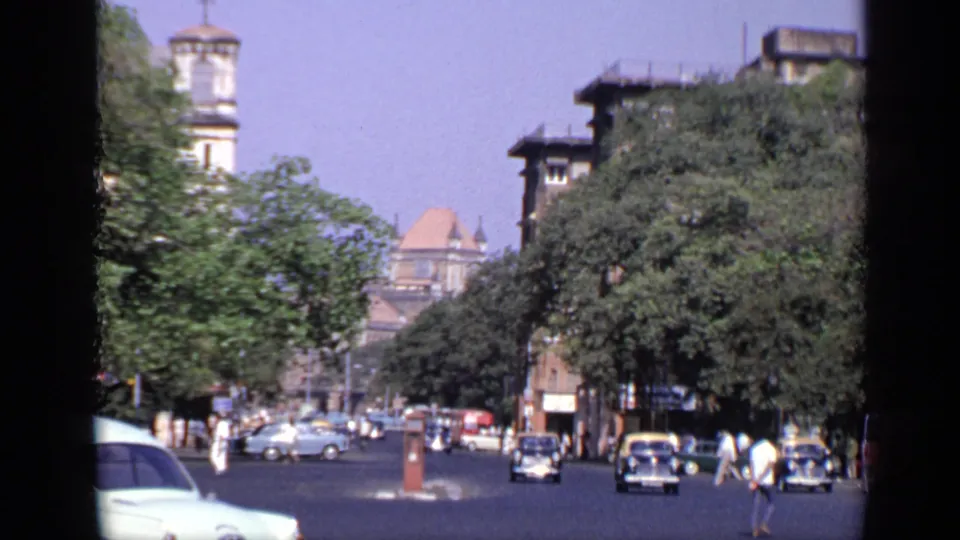1970 India street footage showing cars moving through an open roadway, pedestrians near the curb, trees, and a tower above the street.