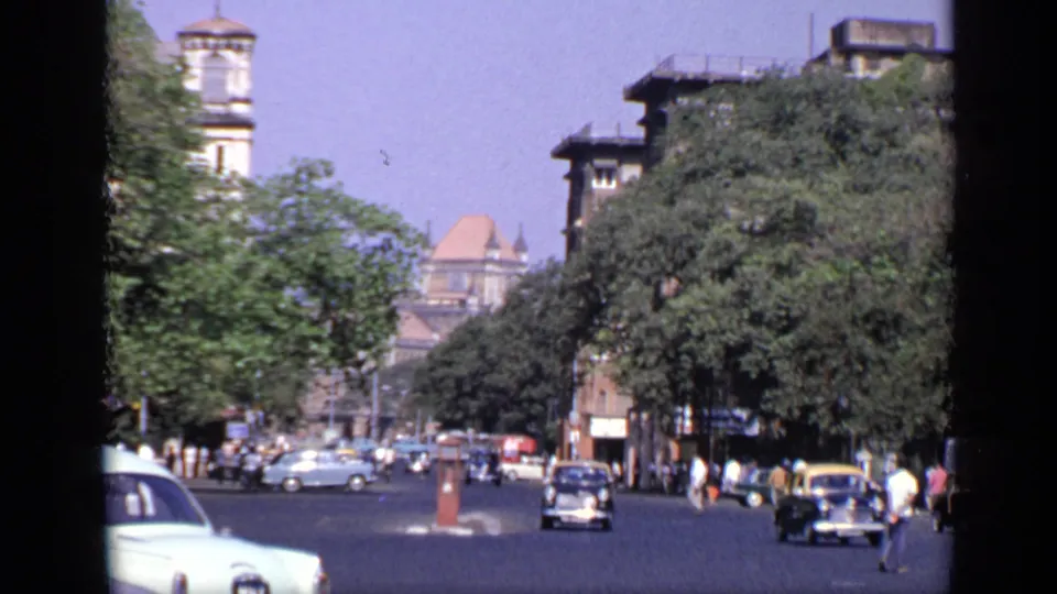 1970 India city street with pedestrians near the roadside, several older cars, dense trees, a visible tower, and colonial-style building details.