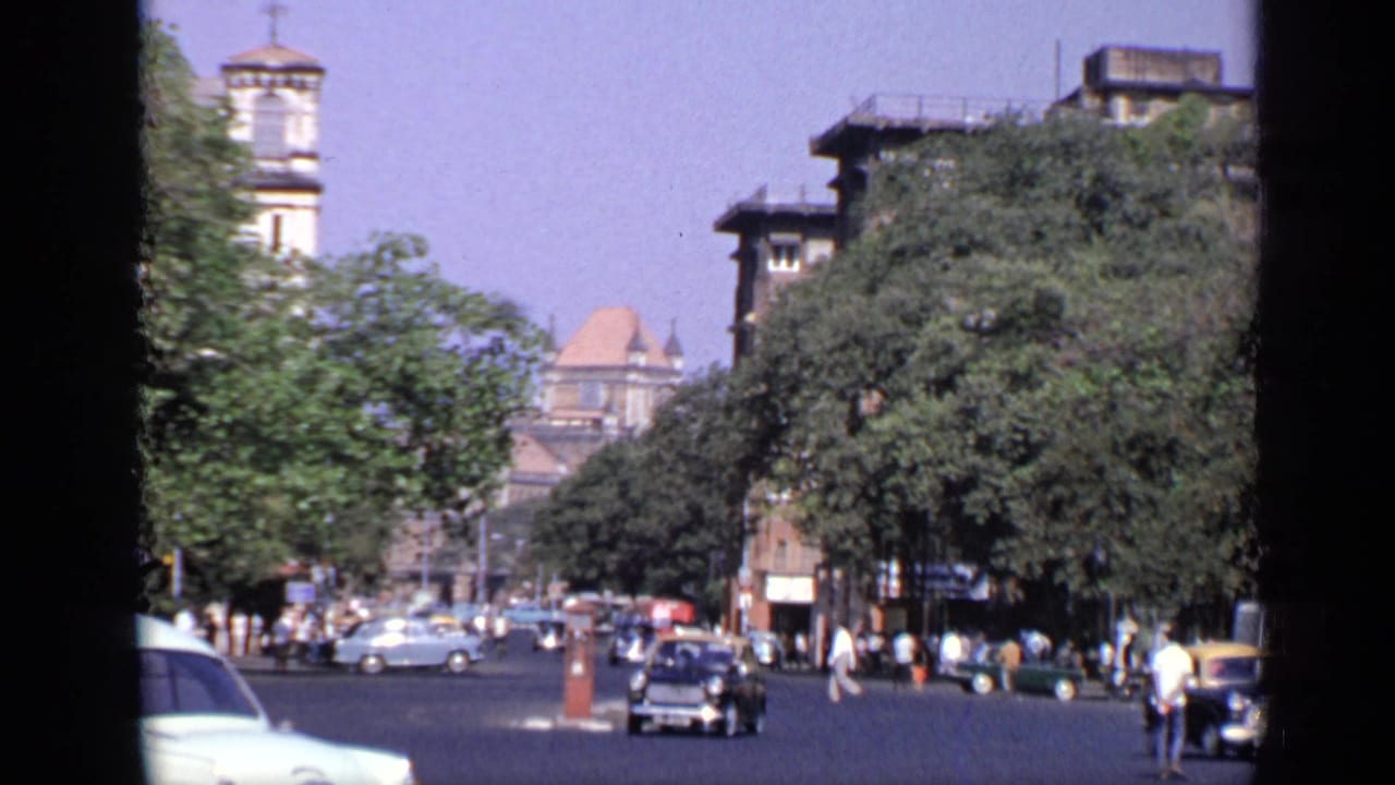 Busy Indian city street with older cars, pedestrians, trees, and large historic buildings.