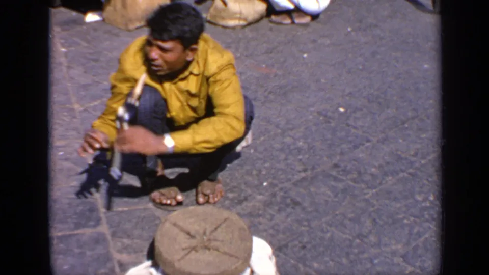 Crouching man in a yellow shirt on an Indian street, handling an object near a round stone piece and sacks.