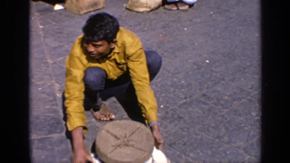 Man in a yellow shirt crouches low and reaches toward a round stone object on the paved street in 1970 India footage.